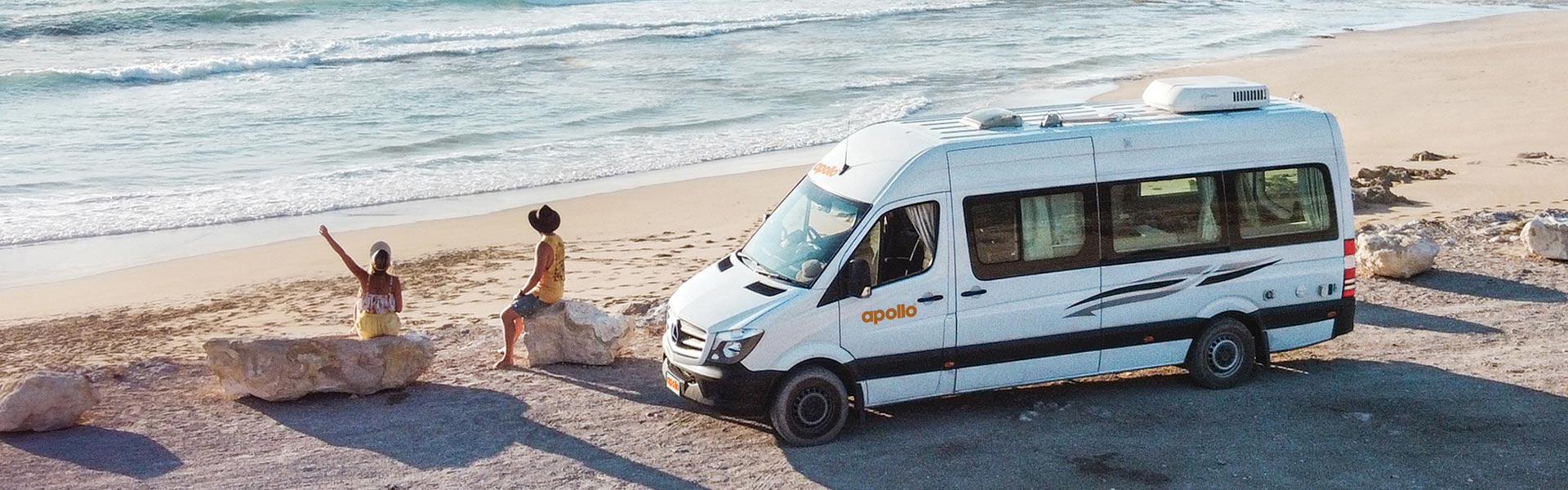 Young man and women sitting on beach in front of Apollo rental campervan